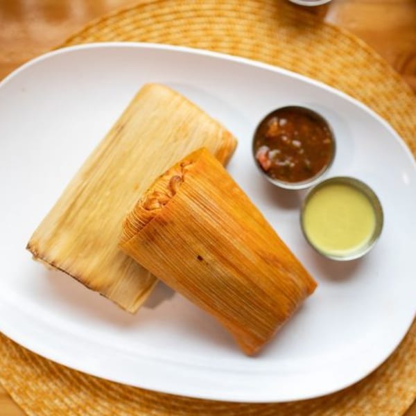 Two tamales, one light and one dark, with two small bowls of salsa on a white plate.