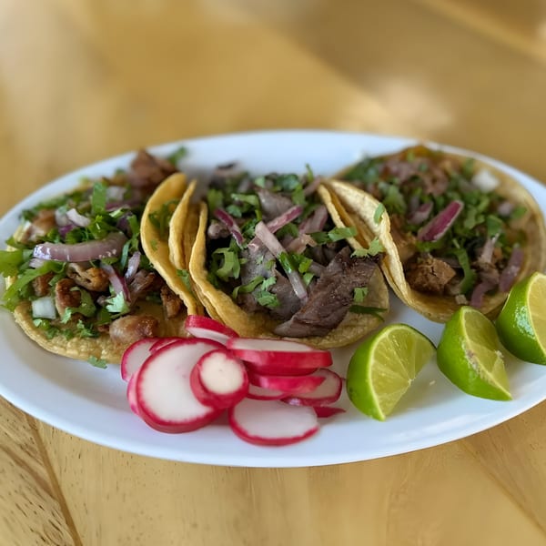 Three Mexican street tacos with meat, cilantro, onions, radishes, and lime wedges on a white plate.