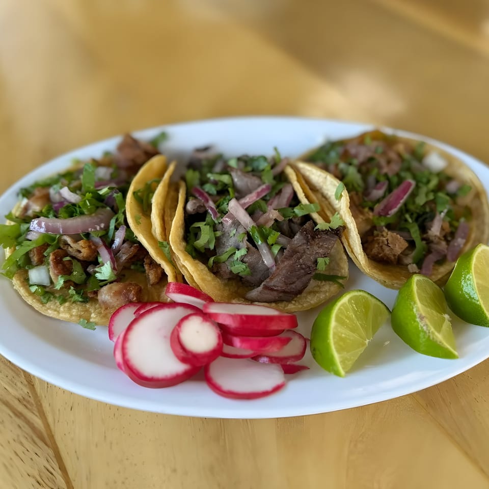 Three Mexican street tacos with meat, cilantro, onions, radishes, and lime wedges on a white plate.