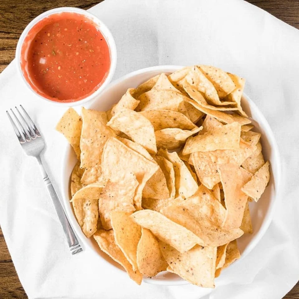 A white bowl of tortilla chips and a small bowl of red salsa on a white napkin.