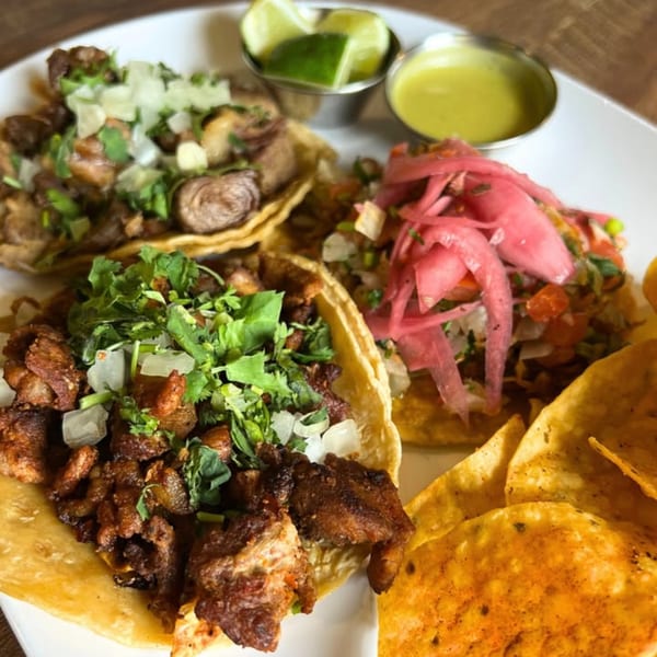 Three Mexican tacos with various toppings, lime wedges, green sauce, and tortilla chips on a white plate.
