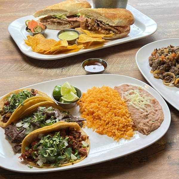 Plates of Mexican food including tacos, rice, beans, tortas, and chips with salsa on a wooden table.